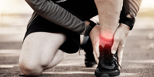 Sportliche Person kniet auf Asphalt und hält sich den schmerzenden Knöchel am rechten Fuß; der Bereich ist rot hervorgehoben, Laufschuh sichtbar.