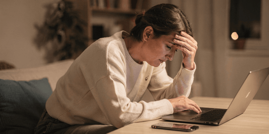 Frau sitzt abends am Tisch vor einem Laptop, stützt die Stirn mit der Hand und wirkt erschöpft; ein Smartphone liegt daneben, warmes Wohnlicht im Hintergrund.