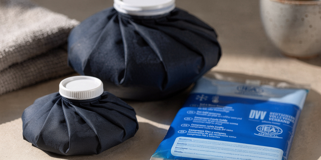 IEA Medical Eisbeutel in zwei Größen neben einer IEA Medical Kalt-Warm-Kompresse auf einem beigen Tisch, mit Handtuch links und einer dampfenden Tasse im Hintergrund.
