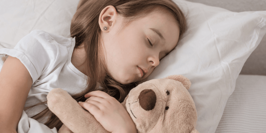 Schlafendes Mädchen liegt im Bett und umarmt einen hellbraunen Teddybären auf einem weißen Kissen.
