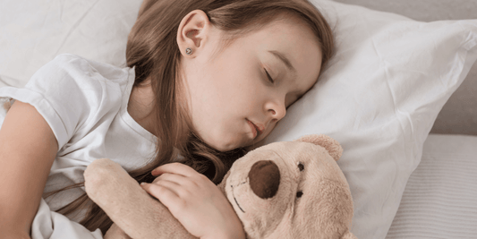 Schlafendes Mädchen liegt im Bett und umarmt einen hellbraunen Teddybären auf einem weißen Kissen.