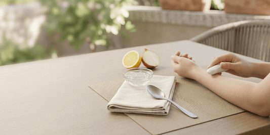 Person hält ein kleines Gerät über dem Unterarm an einem Tisch; daneben Schale mit Wasser, Löffel und halbierte Zitrone.