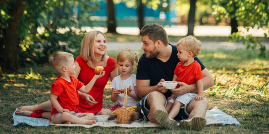 Eltern und drei Kinder sitzen beim Picknick auf einer Decke in einem sonnigen Park mit Korb und Apfel.