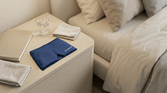 Blauer Stoff Augenmasken Bezug liegt auf einem hellen Nachttisch neben Buch, Wasserglas und gefalteten Tüchern in einem ruhigen Schlafzimmer.