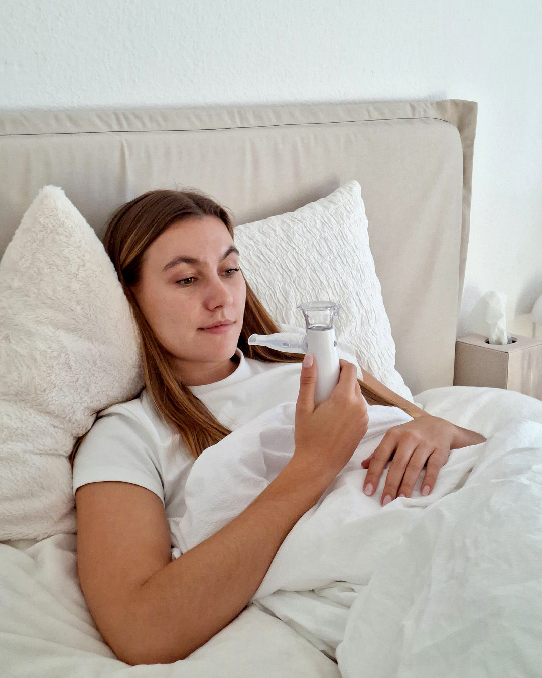 Frau liegt im Bett und hält ein kompaktes Inhaliergerät in der Hand; Anwendungsszene für die Inhalation zu Hause.