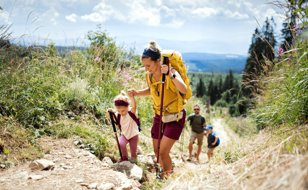 Frau mit gelbem Rucksack und Wanderstöcken führt ein Kind an der Hand über einen Bergpfad, dahinter Mann und Kind.