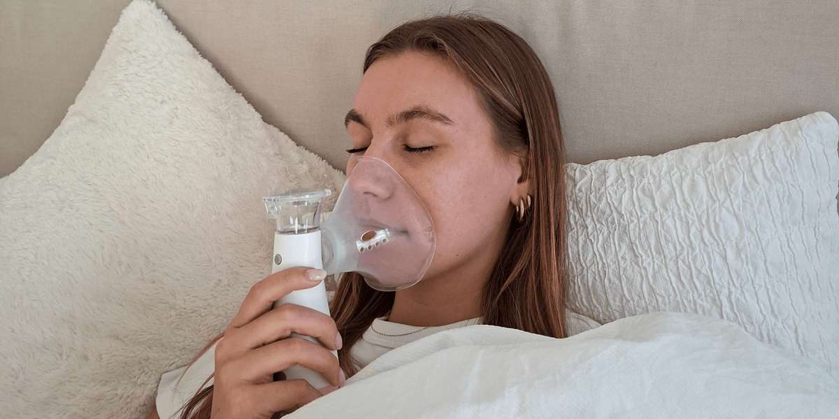 Frau liegt im Bett und inhaliert mit Maske und Handinhalator; Anwendungsszene für die Inhalation zu Hause.