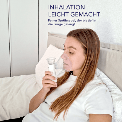 Zurückgelehnte Frau hält einen tragbaren Vernebler mit Maske ans Gesicht, sichtbarer feiner Sprühnebel beim Inhalieren.