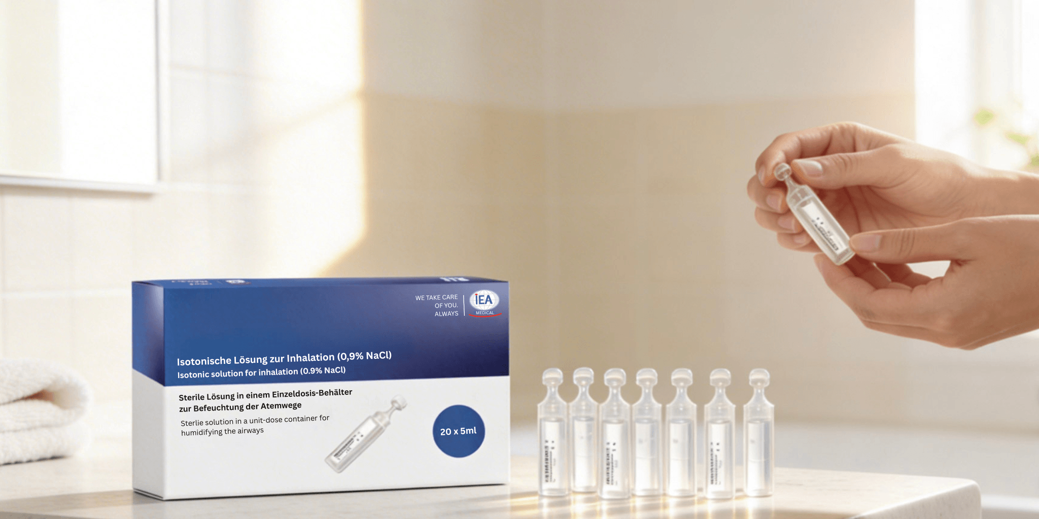 Produktfoto. Die IEA Medical Kochsalzlösung 0,9% NaCl steht auf einer hellen Badeinrichtung. daneben stehen die steril verpackten Einzeldosen. Eine hand öffnet gerade eine Ampulle.