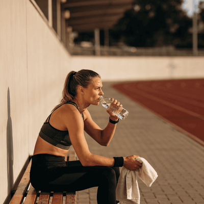 Sportlerin sitzt nach dem Training auf einer Bank und trinkt Wasser, Sportanlage im Hintergrund